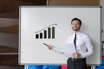 A man is standing in front of a white board with a graph on it. He is holding a piece of paper and...
