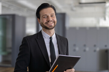 A man in a suit and tie is smiling and holding a black book and a pencil. Concept of professionalism and confidence