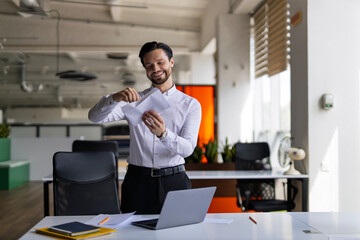 A man in a white shirt is standing in front of a desk with a laptop and a stack of papers. He is smiling and he is in a positive mood. The scene suggests that he is working or studying