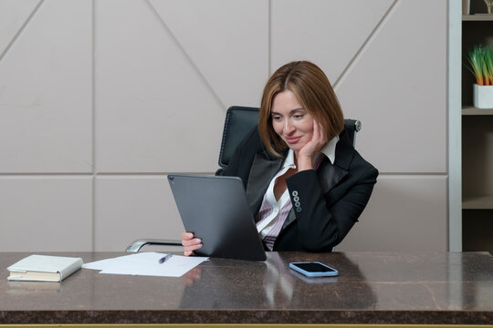 Confident and successful business lady sits at the desk and works on planchet in her modern office	