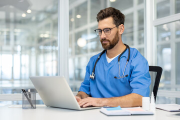 Man doctor in blue scrubs works on laptop in bright, modern medical office. Doctor wearing...