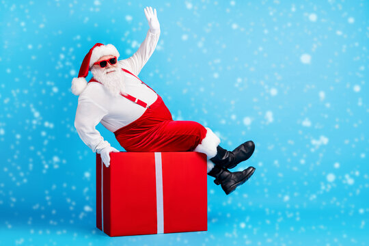 Full length photo of fat overweight santa claus with big stomach beard sit red gift box greeting x-mas christmas party wear suspenders overalls sunglass isolated blue color background