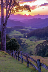 Fototapeta premium Golden hues fill the sky as the sun sets behind mountains, casting a tranquil glow over lush green valleys and a rustic wooden fence