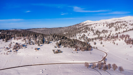 Fototapeta premium An aerial view of a peaceful snow-covered valley with scattered houses, surrounded by dense evergreen forests and rolling hills. The winding road cuts through the pristine winter landscape 