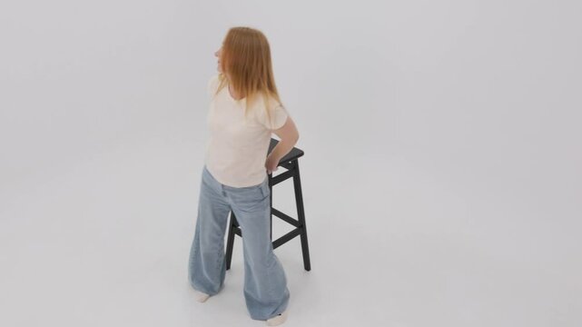 Woman poses and moves gracefully around a stool against a white background in slow motion, showcasing elegance and style