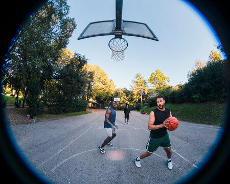 Two basketball players engage in a fierce contest for possession on an outdoor court surrounded by lush trees, showcasing competitive energy and athleticism under soft natural light.