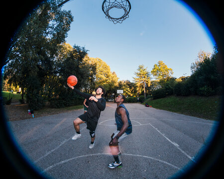 basketball player goes for a layup on an outdoor court surrounded by greenery, highlighting motion and skill with other players in the background under soft sunlight and clear skies