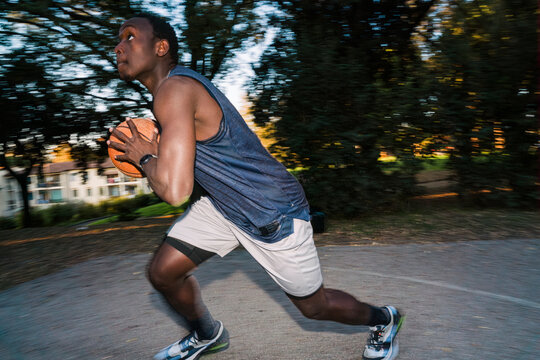 Black young man wearing athletic shorts and a tank top dribbles a basketball on an outdoor court surrounded by trees and nearby residential buildings during a sunny afternoon. flash light motion blur