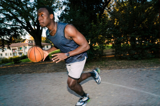 Black young man wearing athletic shorts and a tank top dribbles a basketball on an outdoor court surrounded by trees and nearby residential buildings during a sunny afternoon. flash light motion blur