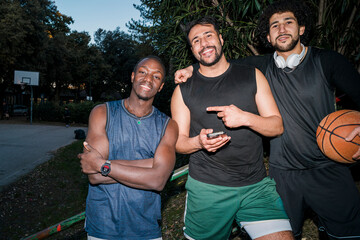 Three young men sitting outdoors on a basketball court playfully taking a selfie, wearing casual sports outfits, laughing and showcasing a strong sense of friendship and fun under twilight skies.