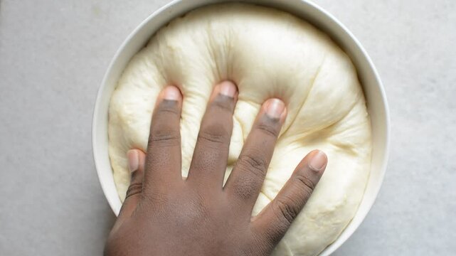 Overhead view of proofed challah dough being deflated, top view of hand deflating dough in a white mixing bowl, process of making challah