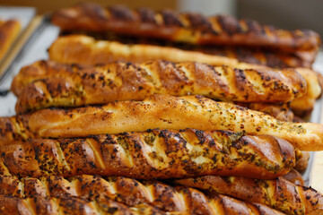 Brown breadsticks, each uniquely textured and topped with herbs, sit enticingly on a tray in a cozy bakery. Freshly baked breadsticks with various toppings resting on a tray in a warm bakery setting