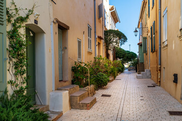 A paved, light-colored alley between ochre-colored houses in St. Tropez with staircases and plants.