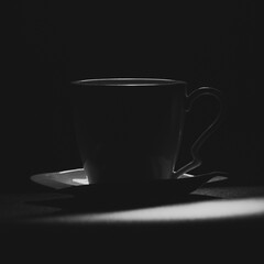A dramatic black and white photograph of a coffee cup with a saucer, captured in low-key lighting. The deep shadows and subtle highlights emphasize the elegant curves and details of the cup, creating 