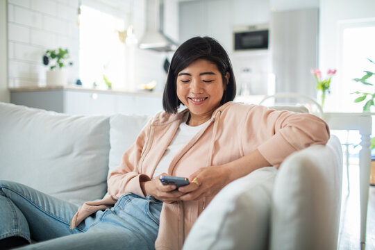 Smiling Asian woman using smartphone on couch at home