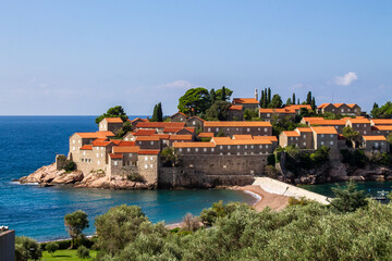 A picturesque coastal village with stone houses topped by orange roofs, surrounded by lush greenery, set against the backdrop of a deep blue sea and clear sky on a sunny day.