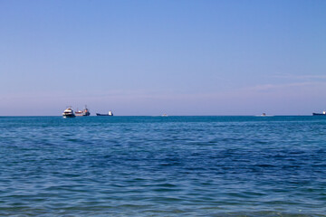 A calm seascape with a clear blue sky, gentle waves, and boats in the distance. The horizon stretches seamlessly, creating a serene and tranquil maritime atmosphere.