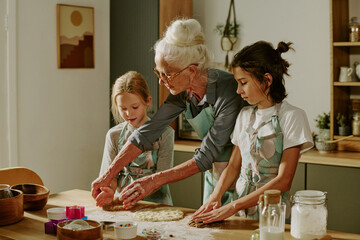 Senior woman guiding two young girls in kitchen while preparing dough together, focused on baking...