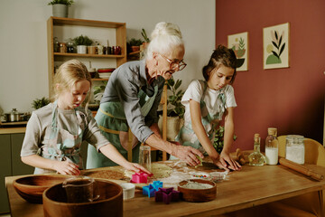 Senior woman teaching two young girls to bake cookies in cozy kitchen with wooden table and various baking utensils. Children focused on cutting out shapes from dough with colorful molds