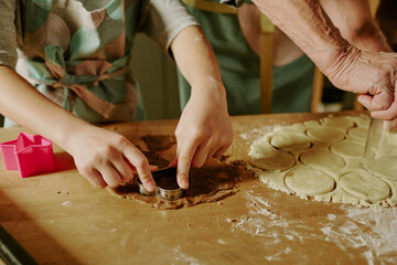 Child and adult working together on dough, cutting shapes using cookie cutter on wooden surface. Hands covered with flour and raw dough cut into various forms