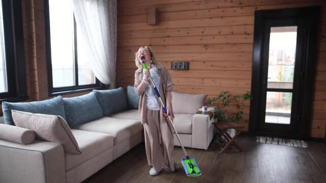 Happy young woman singing while washing floor at living room, using mop as microphone, having fun during house chores. Millennial lady enjoying domestic duties, gamifying cleaning routine