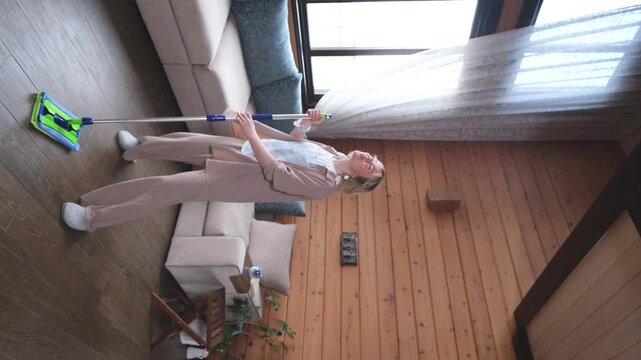 Happy young woman singing while washing floor at living room, using mop as microphone, having fun during house chores. Millennial lady enjoying domestic duties, gamifying cleaning routine