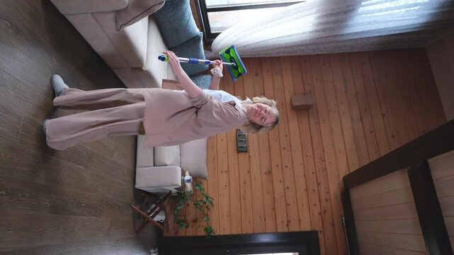Happy young woman singing while washing floor at living room, using mop as microphone, having fun during house chores. Millennial lady enjoying domestic duties, gamifying cleaning routine