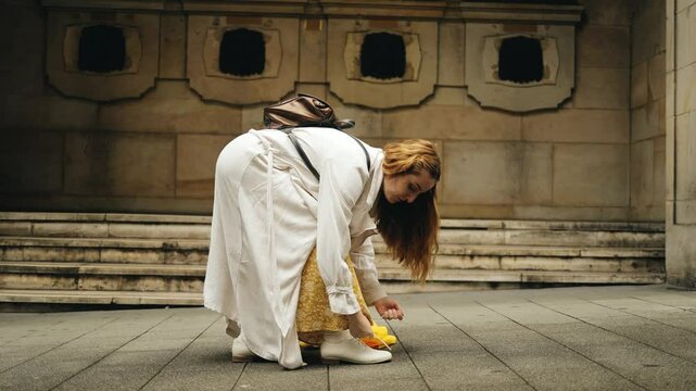 A woman tourist in a historical, Mediterranean city, adjusts her shoe laces in cloudy weather, rain, holding a yellow umbrella in her hands, season, health, cold, tourism, walk, excursion, travel