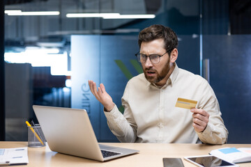 Financial problems. A young sad man is sitting at the desk in the office, holding a credit card in...