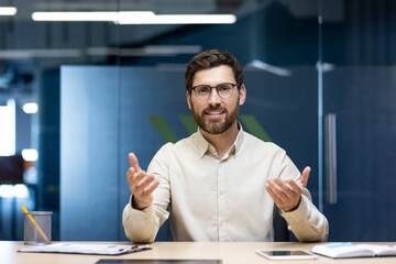 Portrait of a young man sitting in the office at a table in front of the camera and talking on a...