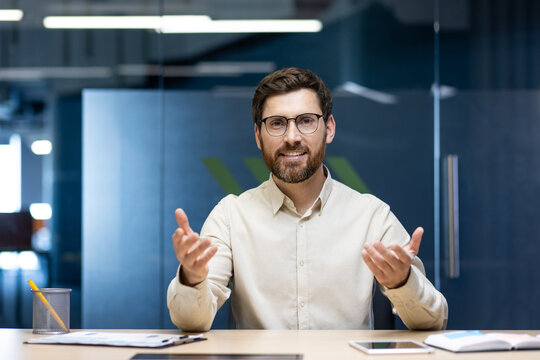 Portrait of a young man sitting in the office at a table in front of the camera and talking on a video call, gesturing to the camera
