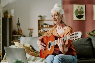 Elderly woman sitting on a sofa, playing a guitar while looking at a laptop in a cozy living room filled with plants and modern decor