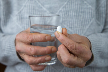 An old woman holds a white pill and a glass of water in her hands.