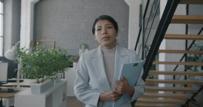 Dolly shot of Middle Eastern businesswoman walking in office holding documents then sitting at desk while people around working. Employee and business concept.