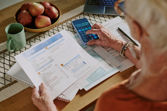 Close-up of elderly person sitting at table, examining financial documents while using smartphone calculator. Visible laptop, green mug, and apples enhance the work environment