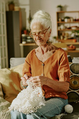 Elderly woman sitting on sofa crocheting in cozy living room filled with plants and shelves in...