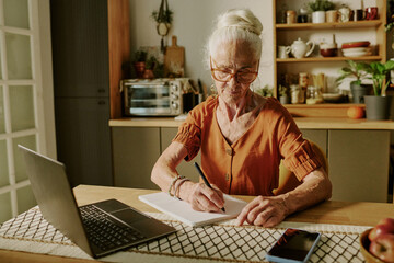 Portrait of senior woman writing on paper while sitting at desk with laptop in cozy kitchen, surrounded by kitchen utensils and decor