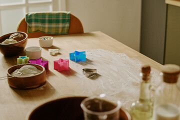 Displaying colorful cookie cutters, bowls, and ingredients spread across wooden table, this image captures joyful anticipation of baking cookies, with sunlight streaming through nearby window