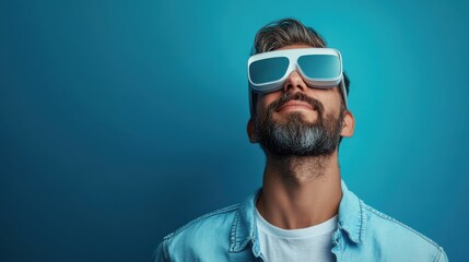 Obraz premium Young man with a beard wearing futuristic sunglasses while looking up against a vibrant blue background, showcasing an imaginative exploration of technology and style.