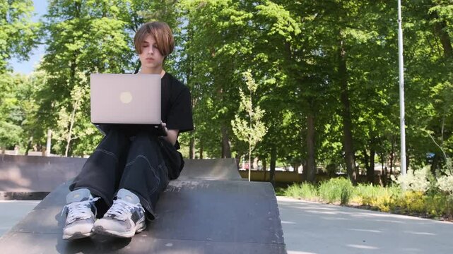 Young man wearing casual clothing uses a laptop while sitting in a skatepark, enjoying a sunny day outdoors