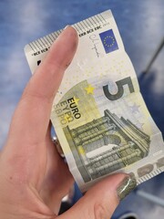 A close-up photo of a five-euro banknote being held in a hand. The banknote shows the iconic European architecture and the signature of the European Central Bank President.