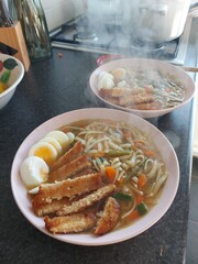 A steaming bowl of homemade ramen with noodles, vegetables, soft-boiled eggs, and crispy fried tofu. The dish is presented on a pink plate and looks both comforting and delicious.
