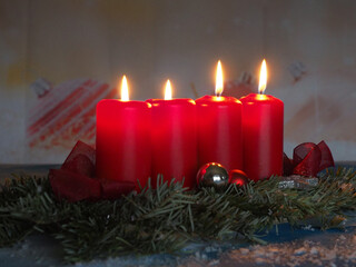 Auf grünen Tannenzweigen stehen vier rote brennende Kerzen zwischen Christbaumkugeln und Schleifen