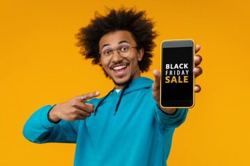 Studio shot of attractive smiling man customer posing over yellow background reaching mobile phone in his hand to the camera and pointing with index finger at its screen with Black Friday interface