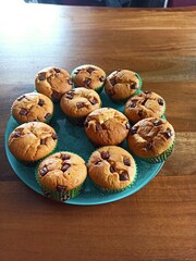 A muffin tray filled with batter and chocolate chunks sits on a kitchen counter, ready to be baked. The baking cups are lined and each has a generous amount of chocolate