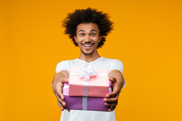 Studio portrait of attractive smiling young African American man reaching the pile of decorated present boxes to the camera. Isolated over yellow background