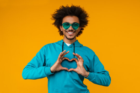 Studio image of attractive smiling young man posing over bright colored orange yellow background wearing trendy eyeglasses and showing heart shape gesture with his hands