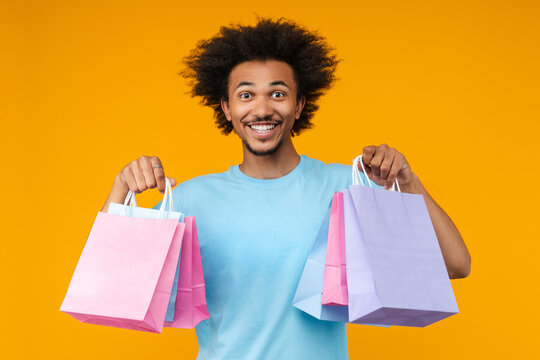 Portrait of excited casually dressed African American young man customer posing in studio with bunches of shopping bags in hands isolated over bright colored orange yellow backgroun