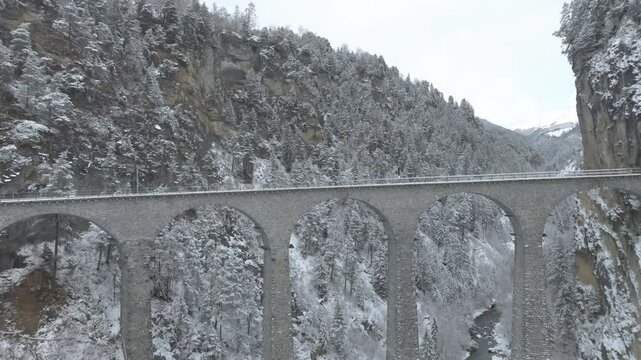 Aerial view of train crossing famous Landwasser Viaduct of RHB narrow gauge railway at Swiss mountain village of Filisur on a snowy autumn day. Movie shot November 22nd, 2024, Filisur, Switzerland.