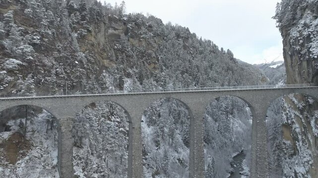 Aerial view of train crossing famous Landwasser Viaduct of RHB narrow gauge railway at Swiss mountain village of Filisur on a snowy autumn day. Movie shot November 22nd, 2024, Filisur, Switzerland.